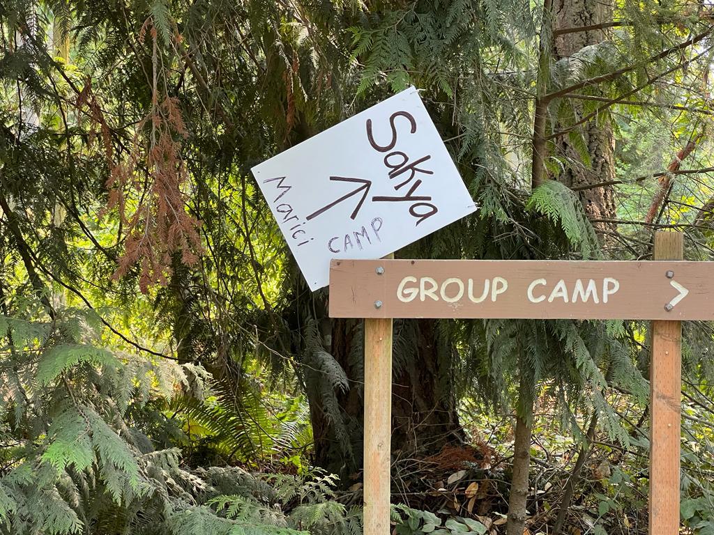 Wooden sign in the forest that says "Group Camp" with a white hand-drawn sign attached at an angle saying "Sakya Marici Camp" and arrow pointing to upper right. 