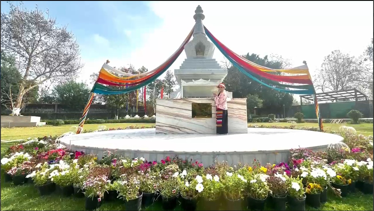 H.E. Dagmo Kushog standing in front of the stupa of H.H. Jigdal Dagchen Dorje Chang, with colorful flowers in foreground and rainbow banners above